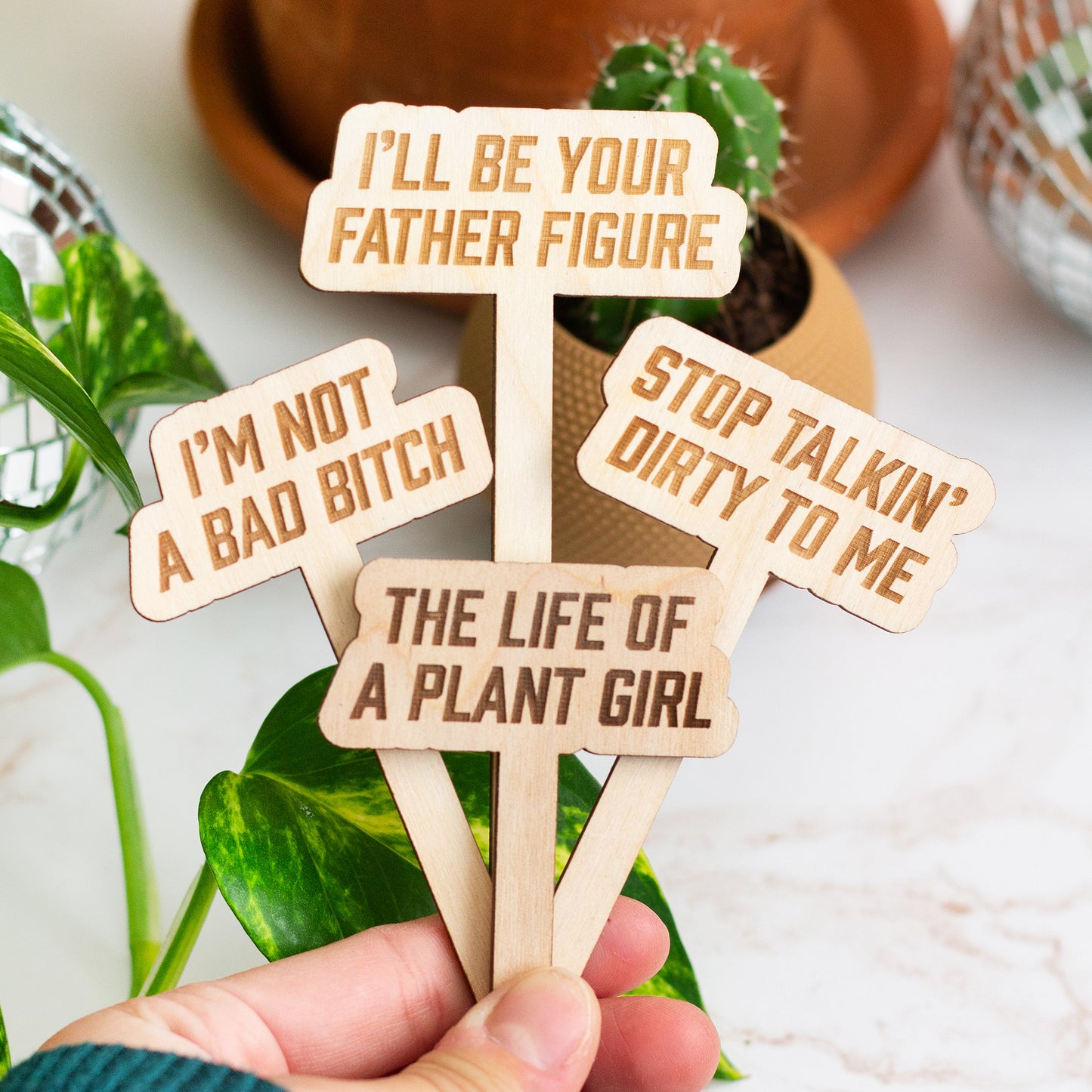 Set of wooden plant labels with humorous text held by a hand, with plants in the background.